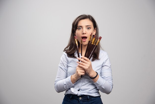 Brunette painter holding bunch of brushes on gray wall. 