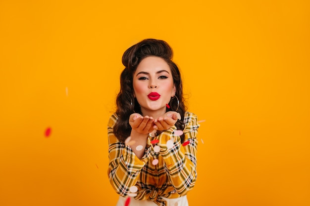 Brunette pinup girl posing with kissing face expression. amazing young lady in checkered shirt standing on yellow background