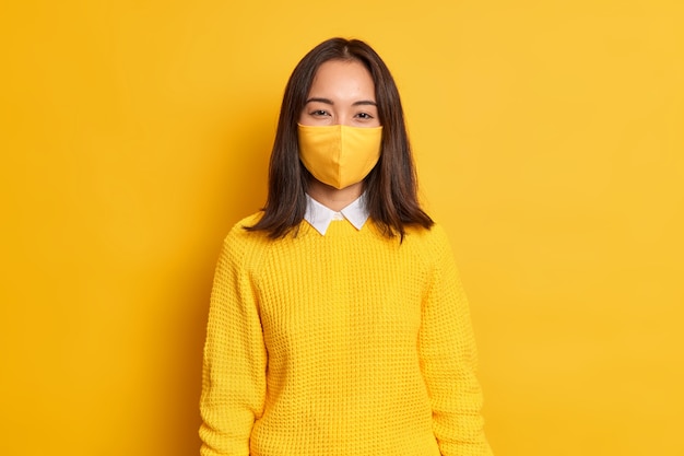 Brunette pretty young asian woman wears protective mask protects herself with coronavirus pandemic dressed in casual jumper