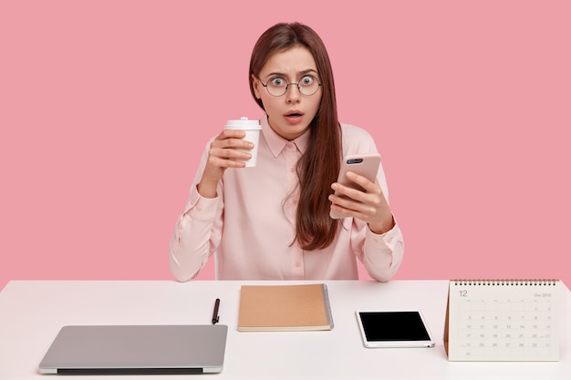 Brunette pretty young female blogger reads recieved notification on cellular, has surprised expression