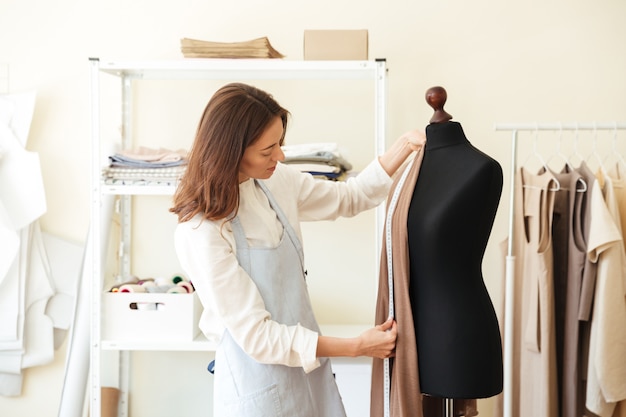 Brunette seamstress in apron measuring beautiful fabric on black dummy in workshop