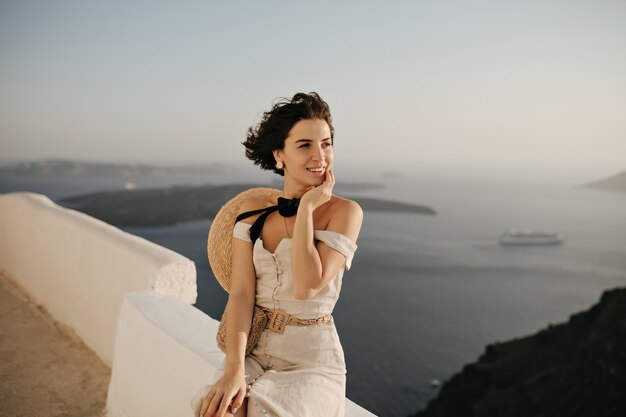 Brunette short-haired woman in elegant beige dress and boater enjoys beautiful sea view