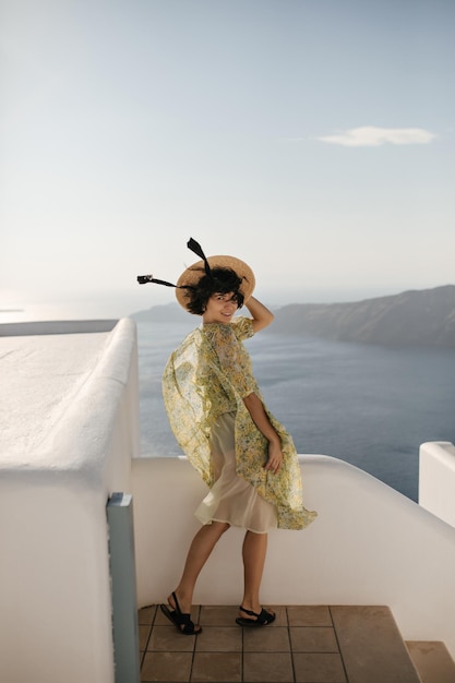 Брюнетка с короткой стрижкой в цветочном стильном наряде улыбается, смотрит в камеру на балконе с видом на море Привлекательная дама в желтом платье держит канотье