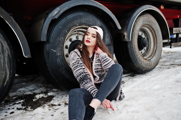 Brunette stylish casual girl in cap sitting against truck wheels