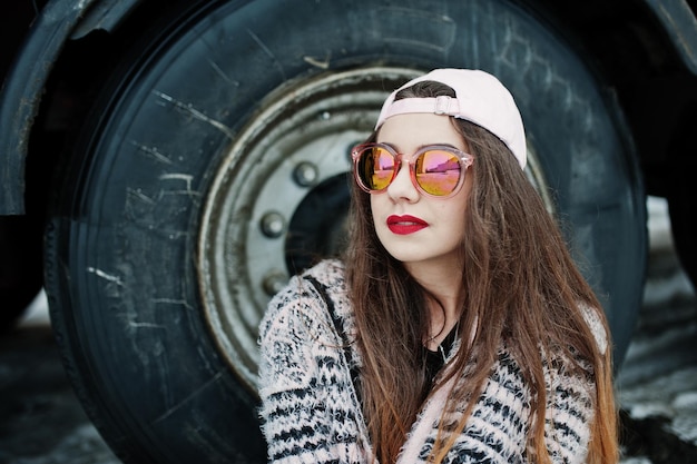 Брюнетка стильная повседневная девушка в кепке и солнцезащитных очках сидит у колес грузовика