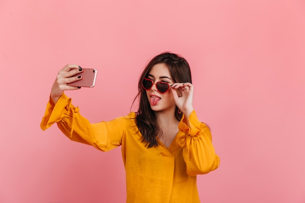 Brunette in sunglasses in shape of heart shows her tongue, making selfie on pink wall.