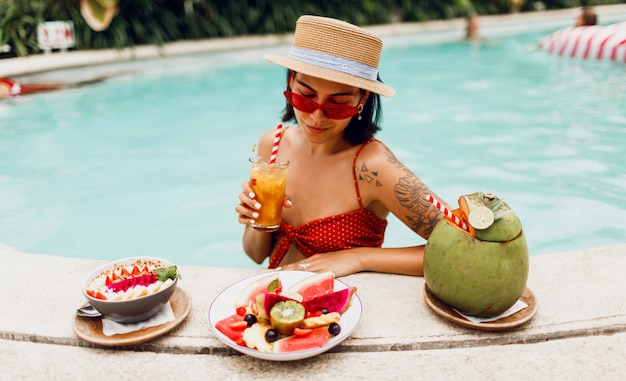 Brunette tan woman in red cat eyes sunglasses  and straw hat relaxing in   pool  with plate of exotic fruits  during tropical vacation.  Stylish tattoo.