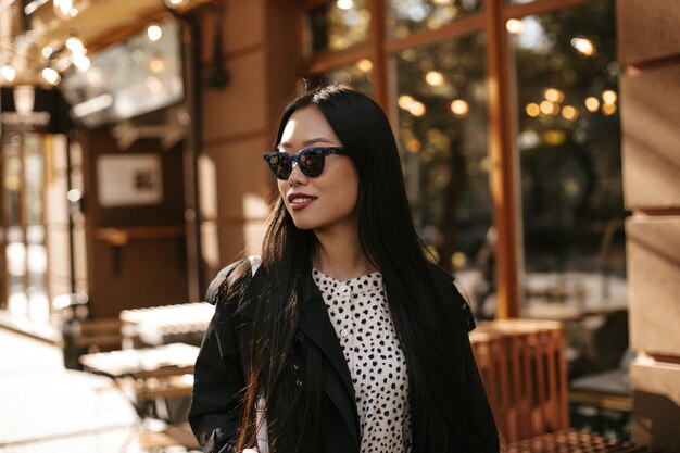 Brunette tanned Asian woman in stylish sunglasses, black trench coat and white blouse smiles