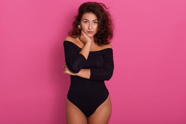 Brunette tanned female with hand touching her neck, having wavy hair. Portrait on rosy, girl wearing black combidress, looks confident.