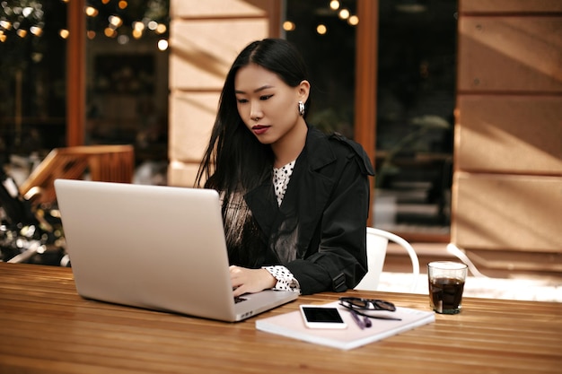 검은색 재킷을 입은 갈색 머리 여자는 컴퓨터 밖에서 작업하고 세련된 옷을 입은 똑똑한 여자는 커피 유리 전화와 노트북이 있는 나무 책상 근처에서 포즈를 취합니다.