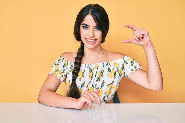 Brunette teenager girl wearing casual clothes sitting on the table smiling and confident gesturing with hand doing small size sign with fingers looking and the camera. measure concept.