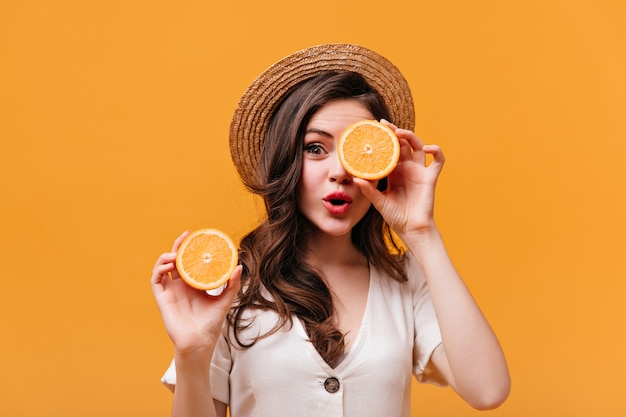 Brunette woman in boater is covering her eye with half orange and looking at camera against isolated background.