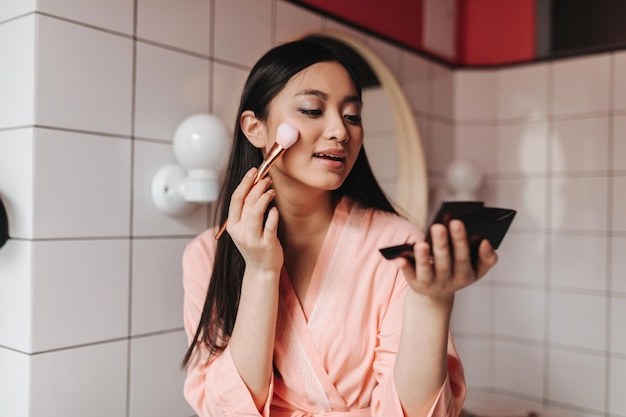 Brunette woman contours while holding small black mirror Lady in pink bathrobe posing in bathroom