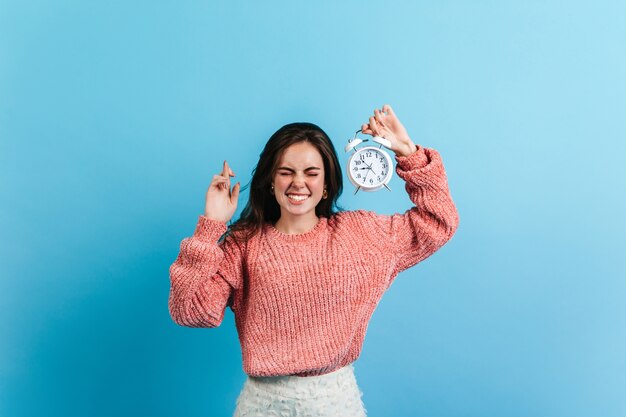 ブルネットの女性は彼女の指を交差させ、白い目覚まし時計を保持しています。青い壁にポーズをとる特大のセーターのモデル。
