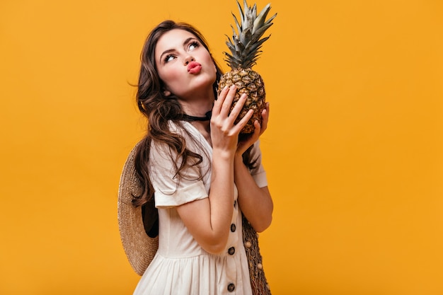 Brunette woman grimaces and holds pineapple on orange background.