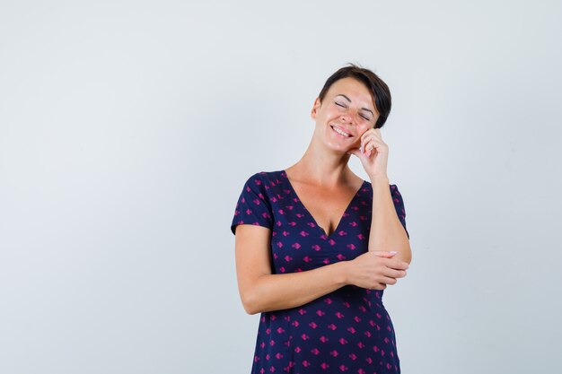 Brunette woman holding her fingers on cheek and closing her eyes in purple and red patterned dress and looking pensive , front view.