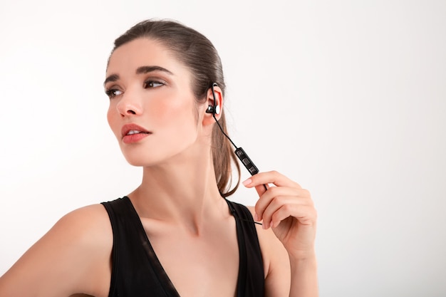 brunette woman in jogging black top listening to music on earphones posing on grey