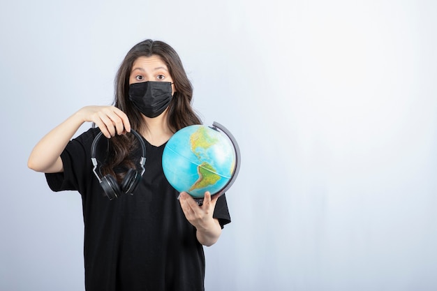 Brunette woman in medical mask holding world globe with headphones . 