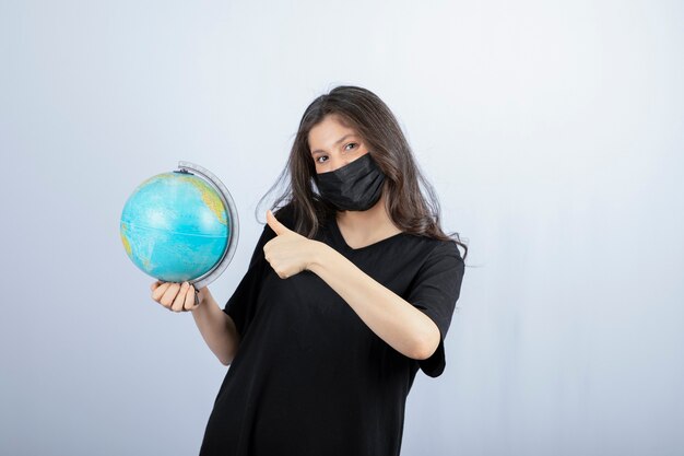 Brunette woman in medical mask with world globe showing thumb up