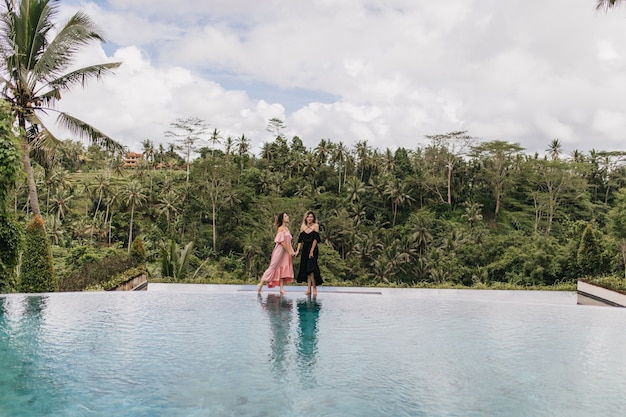 Брюнетка женщина в розовом платье, взявшись за руки с другом на Бали. Наружное фото женских моделей, стоящих возле бассейна в джунглях.