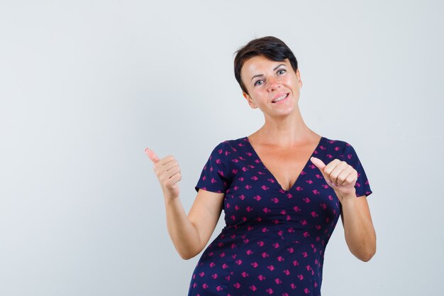 Brunette woman pointing her fingers to the left side in purple and red patterned dress and looking optimistic. front view.