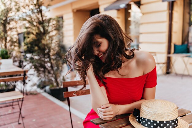 брюнетка женщина позирует с застенчивой улыбкой в открытом ресторане. Портрет веселой кавказской девушки, сидящей за столом в шляпе.