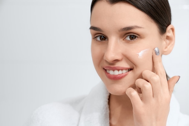 Brunette woman in robe using face cream at home