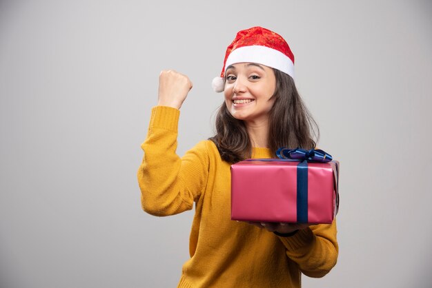 Brunette woman in Santa hat showing her fist and holding gift box. 