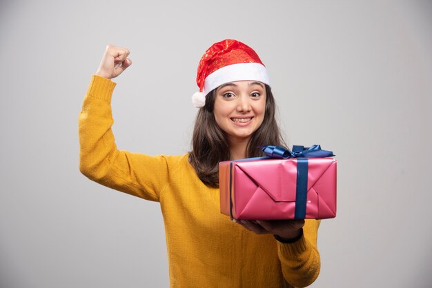 Brunette woman in Santa hat showing her fist and holding gift box. 