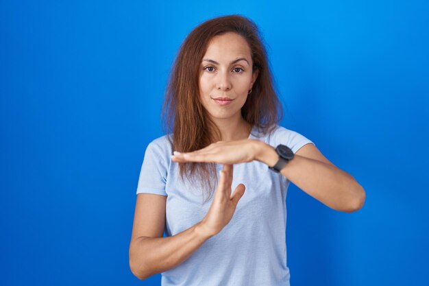 青い背景の上に立って、手でタイムアウトのジェスチャーをし、イライラした真剣な顔のブルネットの女性