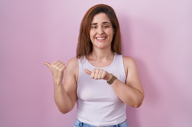 Foto gratuita bruna donna in piedi su sfondo rosa che punta verso la parte posteriore dietro con la mano e il pollice in alto sorridendo fiducioso