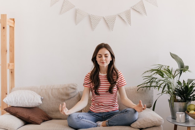 Brunette woman in striped T-shirt is meditating while sitting on sofa in living room