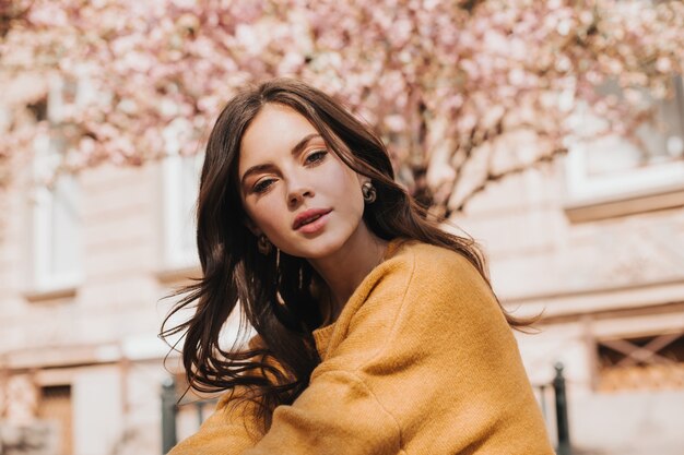 Brunette woman in stylish sweater looks at camera against background of sakura. Lady in yellow outfit posing sensitively outside