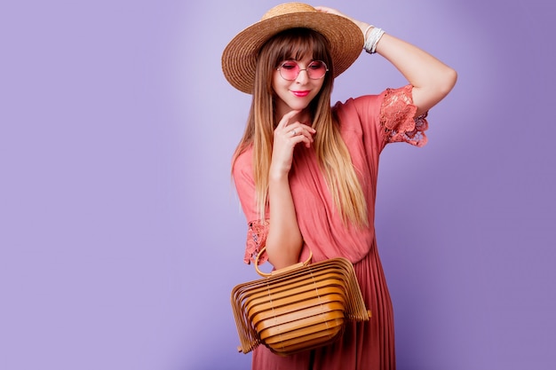 brunette woman in trendy pink dress and straw hat holding bamboo bag on violet.