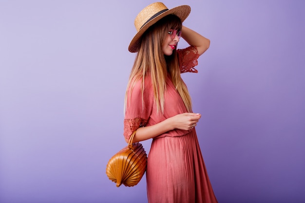 Free photo brunette woman in trendy pink dress and straw hat holding bamboo bag on violet.