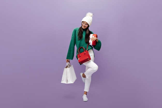 Brunette woman in white pants and green sweater moves on purple background Charming girl in warm hat holds red gift box and shopping bags on isolated