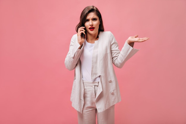 Free photo brunette woman in white suit poses with misunderstanding and talks on phone surprised girl with red lips holds smartphone and looks at camera