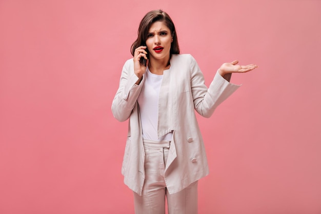 Brunette woman in white suit poses with misunderstanding and talks on phone Surprised girl with red lips holds smartphone and looks at camera