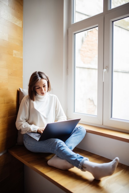 Brunette woman with short hair is working on laptop while sitting on wide windowhill in daily time