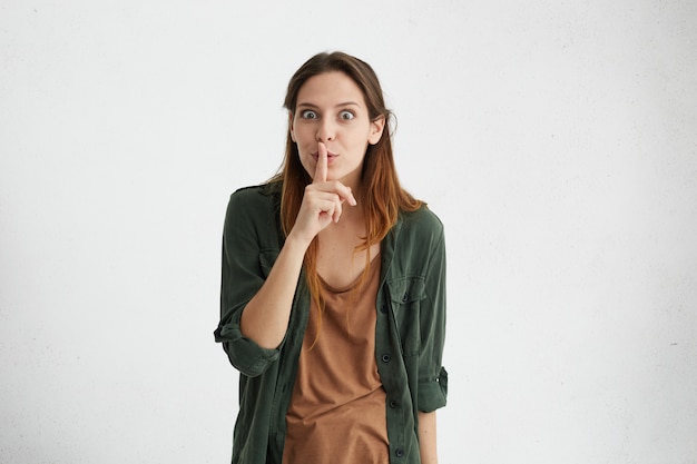 Brunette woman with straight hair wearing green jacket keeping her index finger on lips making hush sign asking not to be noisy.