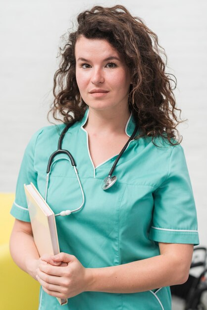 Brunette young female nurse holding book and stethoscope around her neck