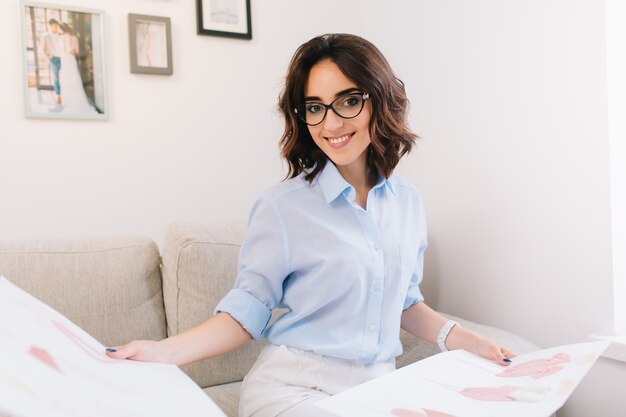 A brunette young girl in a blue shirt is sitting on the sofa in the studio. She has sketches  in her both hands. She is looking to the camera.