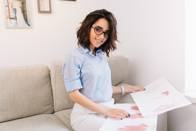 A brunette young girl in a blue shirt is sitting on the sofa in the studio. She has sketches  on her knees. She is looking to the camera.