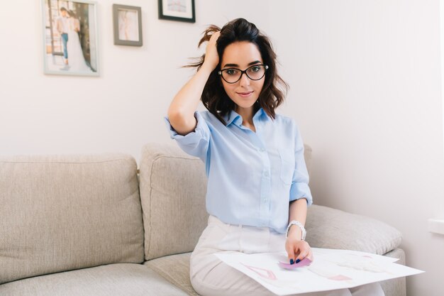 A brunette young girl is sitting on the sofa in studio. She wears blue shirt and white pants. She has sketches and some material on her knees.