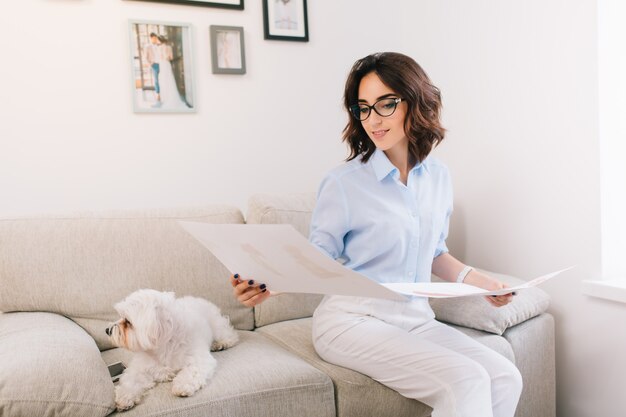 A brunette young girl is sitting on the sofa in studio. She wears blue shirt and white pants. She is looking at the sketches which are in her both hands.