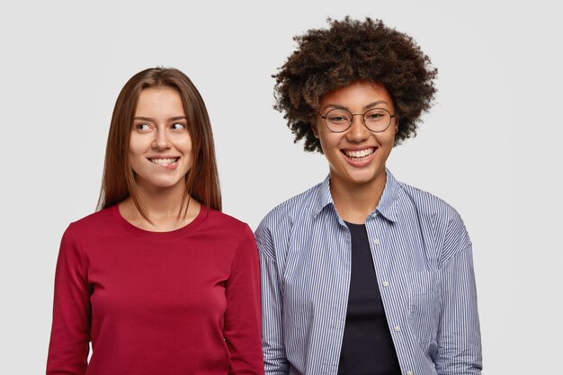 Brunette young woman has intention to do something, bites lower lip, looks with happiness aside, stands near dark skinned female companion, have joy together, isolated over white  wall