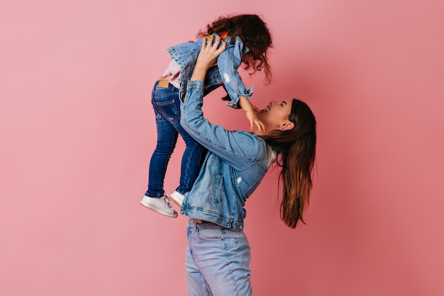 Brunette young woman holding daughter on pink background. Studio shot of mom and preteen kid in denim jackets.