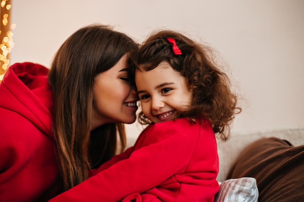 Brunette young woman kissing child. Indoor shot of mom and little kid smiling at home.