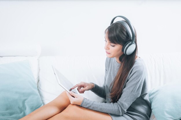 디지털 태블릿으로 음악을 듣고 소파에 앉아 휴식을 취하는 갈색 머리 젊은 여성