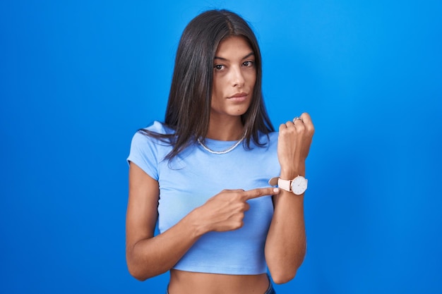 Brunette young woman standing over blue background in hurry pointing to watch time impatience looking at the camera with relaxed expression